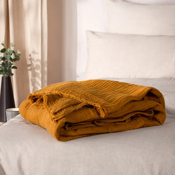Folded mustard yellow blanket on a bed with white pillows and curtains in the background.