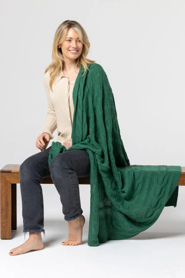 Woman sitting on a bench with a green blanket draped over her, wearing a beige cardigan and dark jeans.