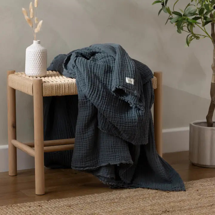 Blue textured blanket draped over a wooden stool with a white vase and plant in the background.