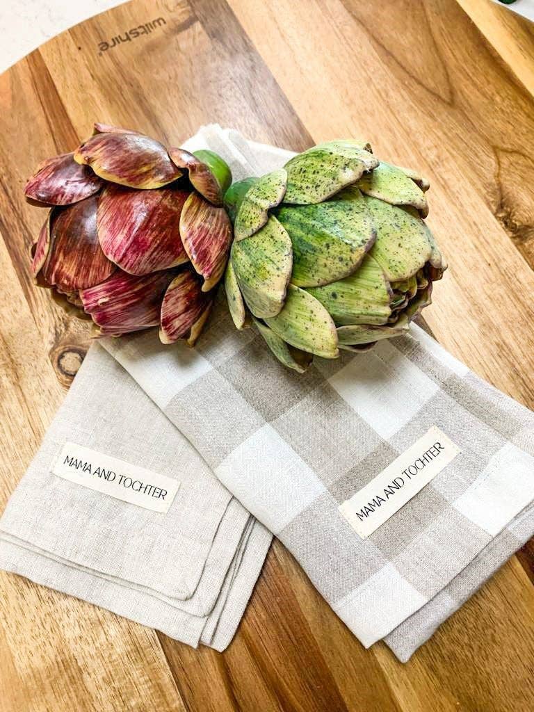 Two artichokes on a wooden surface with checkered towels underneath, branded 'Mama and Tochter'.